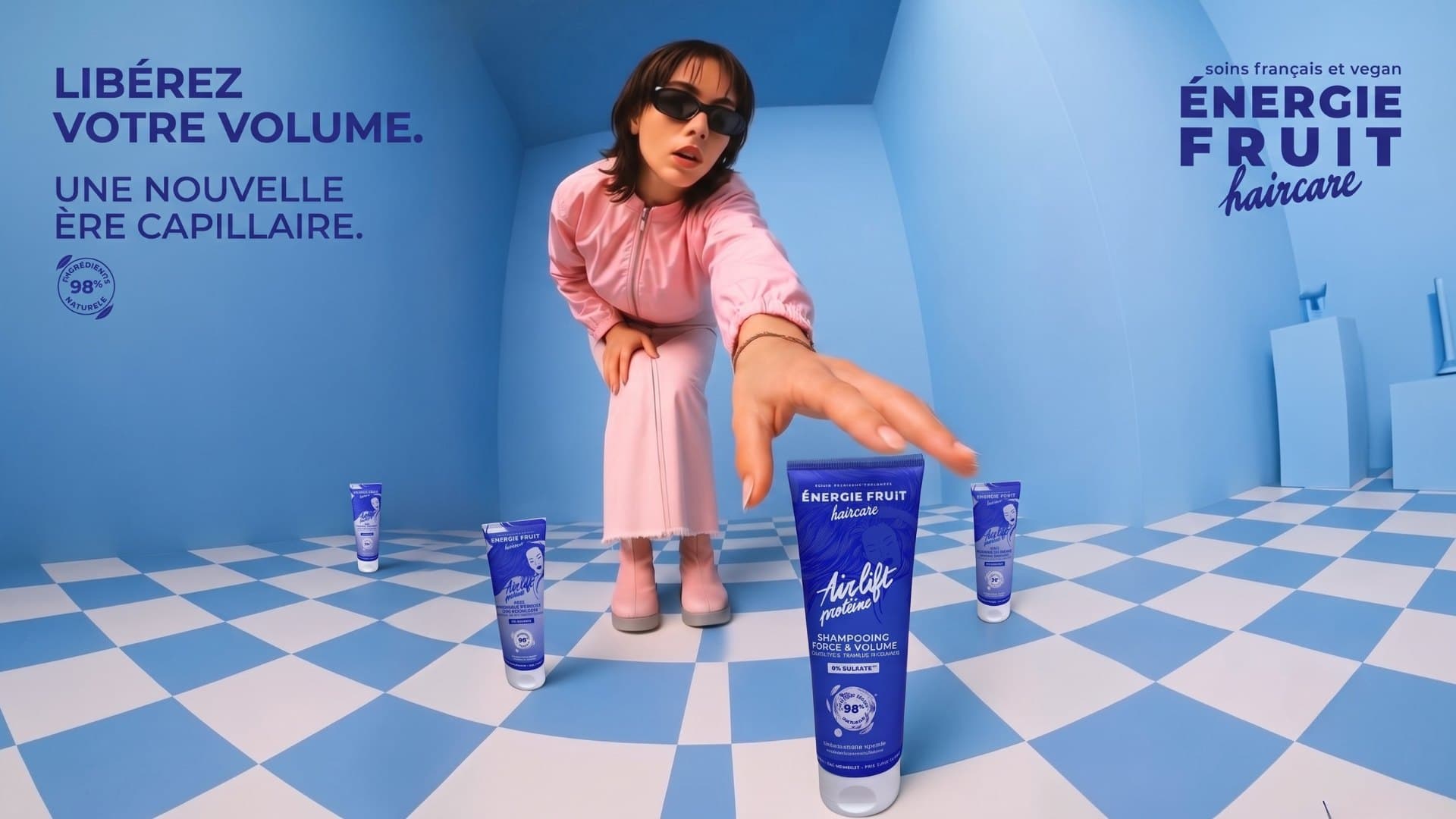 Libérez votre volume — une nouvelle ère capillaire. Un mannequin tend la main vers un tube de shampooing dans une pièce bleue.